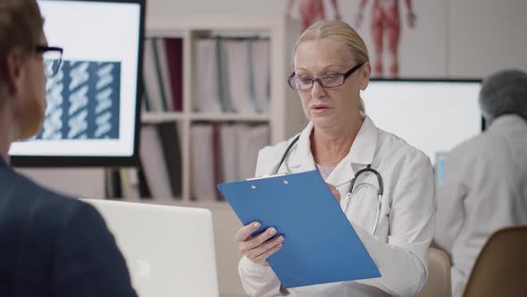 Mature Female Doctor with Clipboard Talking to Young Male Patient in Hospital Office