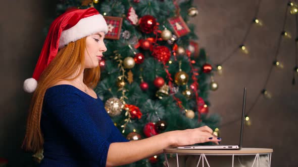 Woman in Santa Hat Works Laptop and Types on Keyboard Background Christmas Tree.