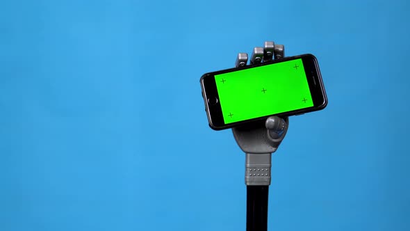 A Mechanical Hand Holds a Phone with a Green Screen. Gray Cyborg Hand Holding a Smartphone on a Blue