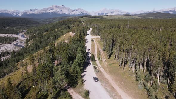 Multiple cars driving along a dusty logging road through a boreal forest with a majestic Rocky Mount