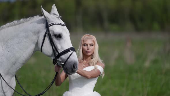 Beautiful Blonde Woman in Wedding Dress and White Horse are Walking at Nature