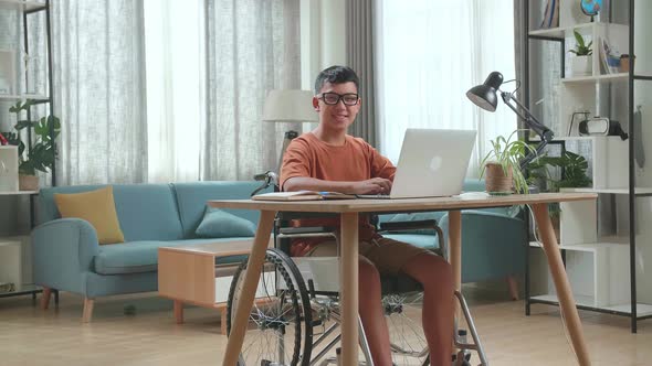 Asian Boy Sitting In A Wheelchair Using Laptop Computer While Looking And Smiling To Camera At Home