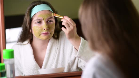 4k Smiling young woman applying clay facial mask looking in bathroom mirror