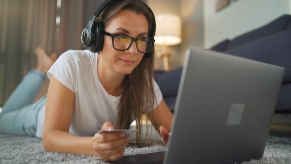 Woman with Glasses and Headphones Is Lying on the Carpet, Listening To Music and Makes an Online