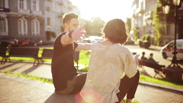 Young Beautiful Couple Smiling Speaking Looking at Tablet Sitting in City Park