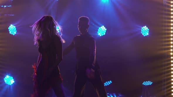 Passionate Blonde is Dancing Rumba with Her Partner Against Backdrop of Colorful Lights
