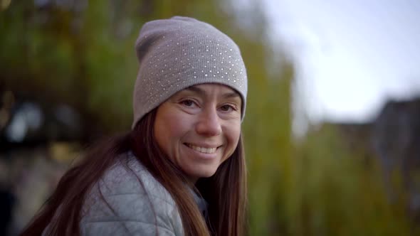 Portrait of a Cute Brunette with Long Hair and a Gray Hat on a Background of Green Trees. She Looks