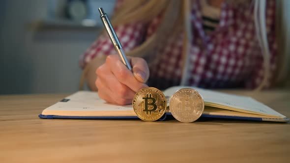 Woman Writing in Notebook with Bitcoins Nearby. Crop View of Female Hands in Checkered Shirt