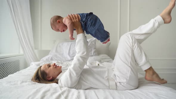 Happy Healthy Young Adult Caucasian Mum Lifting Cute Infant Child Up Playing Airplane on Bed