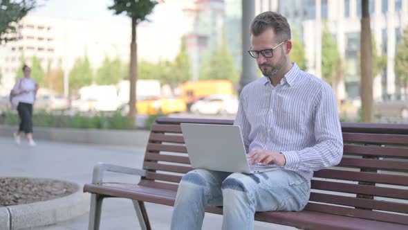 Young Adult Man with Laptop Looking at Camera While Sitting Outdoor on Bench