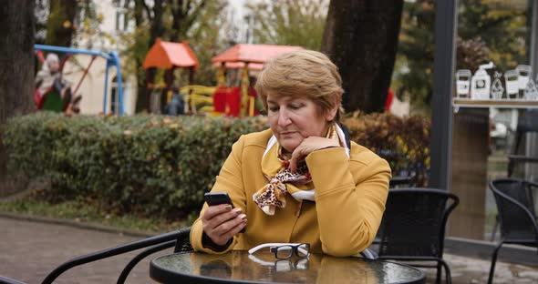 Senior Attractive Woman Sitting on a Chair Near an Outdoor Cafe Uses Her Phone on a Social Network