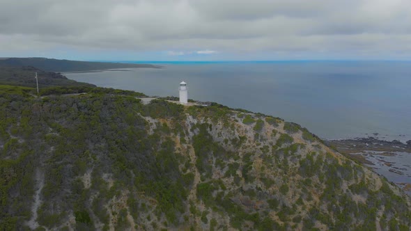 Aerial of a lonely white lighthouse sits on top of a high cliff overlooking the ocean.