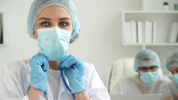 Female Doctor Corrects Mask on Background of Colleagues at Computer in Clinic