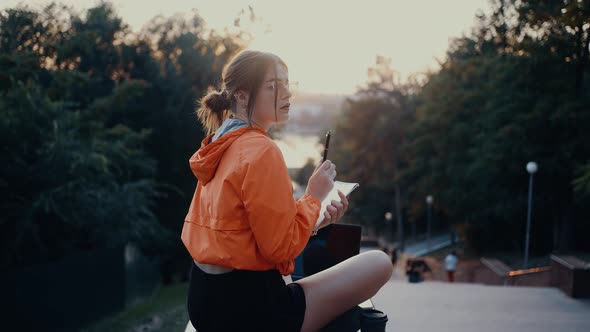 A Creative Young Woman in Glasses Makes Notes in the Notebook in Her Hand Sitting on the Bench