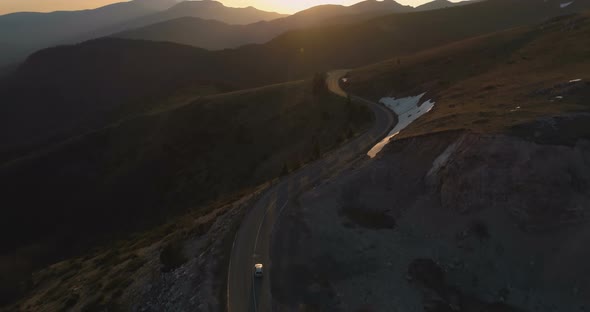 White Car Driving on Winding Remote Mountain Road. Sunset Lights Shine Over Mountain Peaks