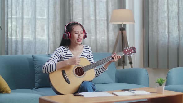 Asian Woman Composer Wearing Headphones Singing And Playing Guitar At Home