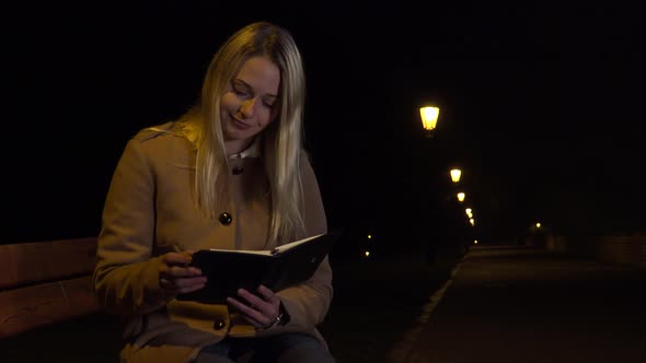 A Young Beautiful Woman Sits on a Bench and Writes Into a Notebook in an Urban Area at Night