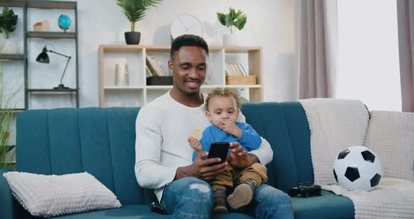 Father which Holding His Small Son on Knees and Uses His Mobile, Sitting on Soft Couch at Home