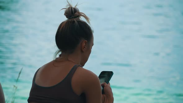 Young Woman in a Cap Sits Near the River in a Smartphone in Nature