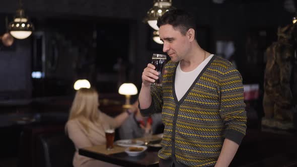 Happy Satisfied Man Smelling Drinking Dark Lager Standing in Restaurant with Blurred Family at