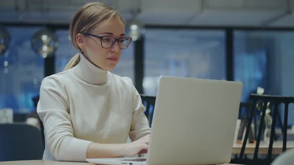 Young Woman in Glasses Works on a Laptop While Sitting in an Evening Cafe