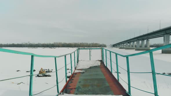 Green Rusty Pier Stands at Frozen River Against Large Bridge