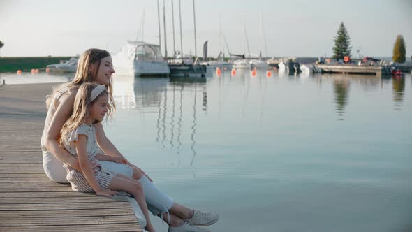 Woman with Daughter Sit on a Pier Looking at Lake at the Sunset