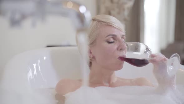 Charming Woman Drinking Red Wine in Bathtub. Portrait of Young Blond Caucasian Lady with Grey Eyes