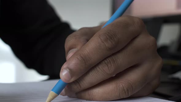 Left Hand Of Ethnic Minority Male Writing With Blue Pencil. Low Angle, Close Up, Locked Off