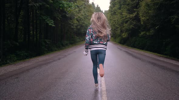 Slow motion shot of a blonde girl running in the middle of a road surrounded by woodland. Dolly Push