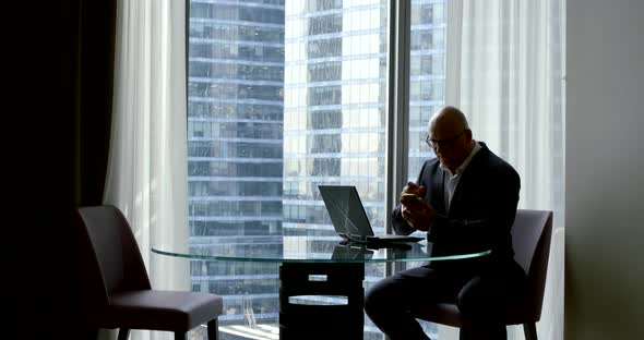 Adult Rich Man Is Sitting at Table in His Apartment and Typing on Smartphone, Open Laptop