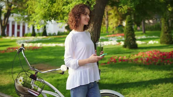 Happy Smiling Woman Messaging Using Her Smartphone Standing in the City Park Near Her City Bicycle