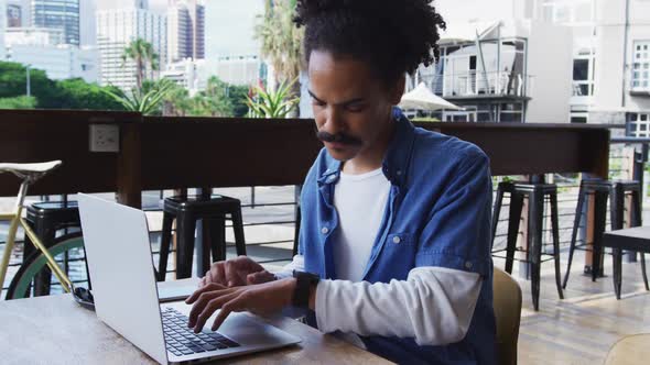 Mixed race man with moustache sitting at table outside cafe using laptop and smartwatch