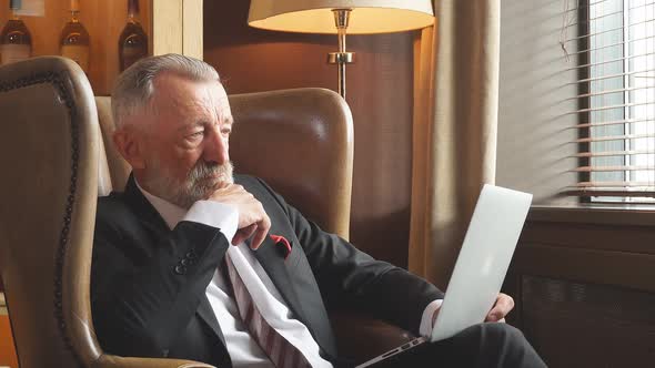 Portrait of Old Man with a Calm Face, Sitting in Dark Office, Keeps Laptop Computer and Reading News