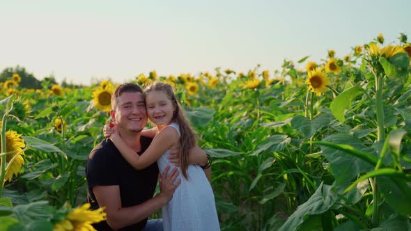 Father and daughter hug looking at camera at sunflowers field on sunset
