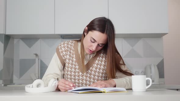 Young Woman with Expression Reads a Book While Sitting at a Table in the Kitchen