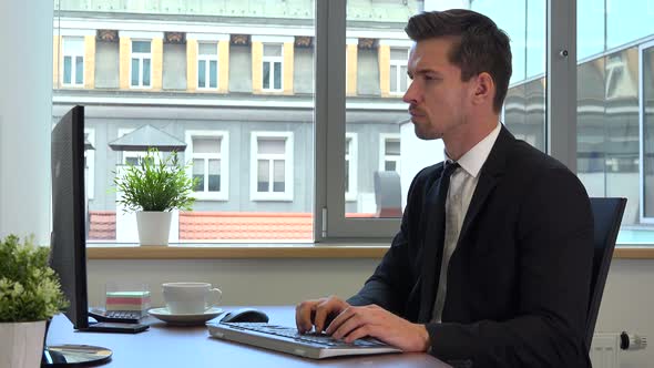 An office worker in a suit works on a computer, looks seriously at the camera