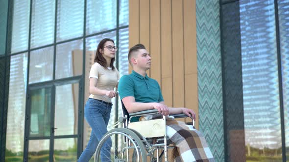 A Young Woman Is Carrying a Young Man in a Wheelchair. An Assistant Carries a Disabled Person in a