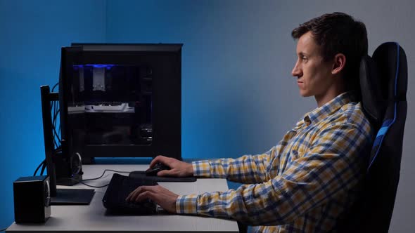 Man Plays Videogame on Large Computer Screen Sitting in Gaming Chair in Semidark Room Illuminated By