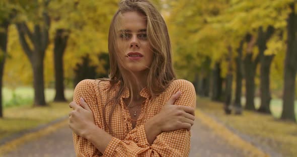 Freezed Caucasian Girl with Brown Hair and Pleasant Smile Standing on the Road in the Autumn Park