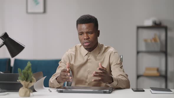 Young Black Man is Sitting at Table in Room Talking and Gesticulating Looking at Camera
