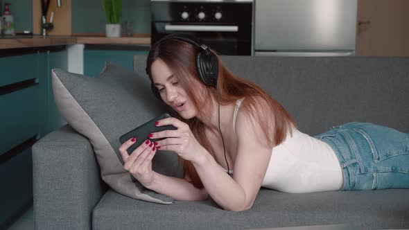 Young Woman is Playing Video Game in Smartphone By Internet Using Headphones and Lying on the Couch