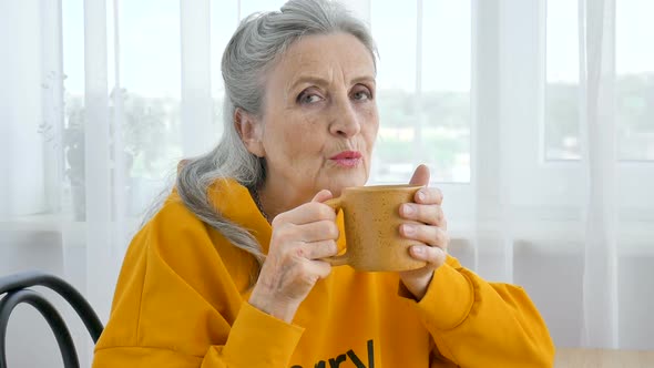 Happy Elderly Pensioner Eccentric Grayhaired Woman in Yellow is Drinking Tea Looking Thoughtfully