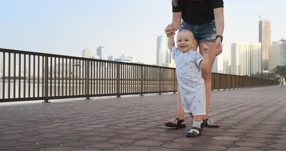 Smiling Boy Holding His Mother's Hand Makes the First Steps Walking Along the Promenade in the