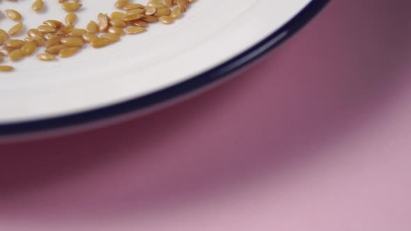 Flax seeds fall in a heap on a white plate with a blue border on a pink bright background.