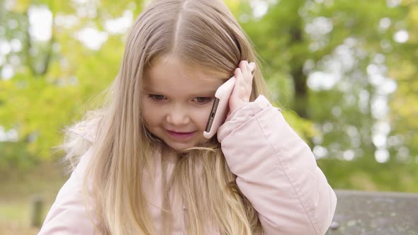 Cute Little Caucasian Girl Talks on a Smartphone with a Smile As She Sits on a Bench in a Park