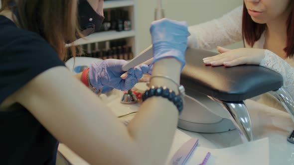 Unrecognizable Person in Gloves Saws Nails of Young Woman in Salon