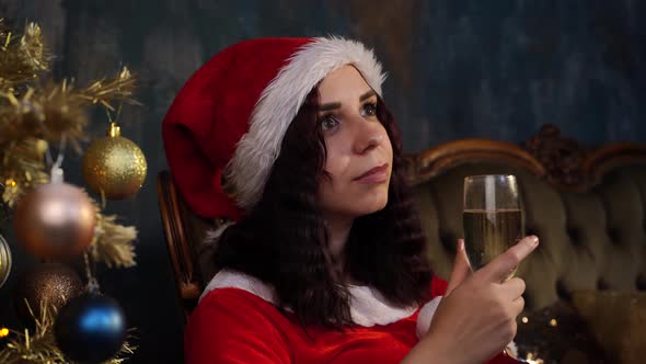 Young Thoughtful Woman in Santa Claus Costume with Glass of Champagne Sitting on Chair Near