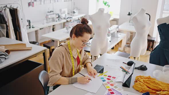 Female Fashion Stylist Sitting with Digital Tablet at Workshop Drawing Sketches of New Collection