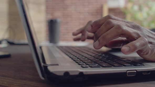 Close-up of the hands of a black man typing fingers on laptop keyboard at an office table.